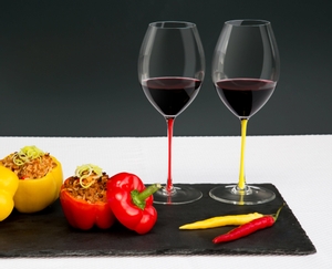 Two red wine filled RIEDEL Fatto A Mano Syrah glasses with a red and a yellow stem on a slate plate with stuffed peppers.