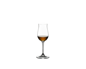 A RIEDEL Vinum Cognac Hennessy glass filled with cognac stands on a table between a typewriter and three open cognac bottles. The sun shines through the window into the room and makes the cognac in the glass shine.