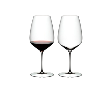 Two RIEDEL Veloce Cabernet/Merlot glasses one filled with red wine and an unfilled glass on a white background.