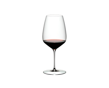 Two RIEDEL Veloce Cabernet/Merlot glasses one filled with red wine and an unfilled glass on a white background.