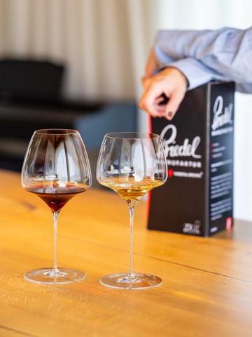 Two RIEDEL wine glasses filled with red and white wine on a wooden table, with a RIEDEL Manufaktur box and a person’s hand in the background, symbolizing elegance and craftsmanship.