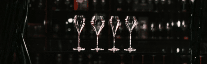 Four elegant RIEDEL wine glasses stand in a row on a reflective black surface, illuminated softly against a dark background showcasing subtle reflections of fine glassware.