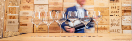A row of six RIEDEL wine glasses displayed on a wooden table, each with a unique bowl shape, set against a backdrop of wooden wine crates with a blurred figure in the background.