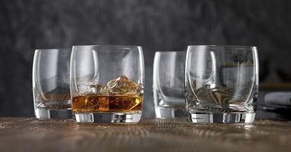 Four NACHTMANN Vivendi whiskey tumblers on a wooden table, one of them is filled with whiskey on the rocks.<br/>