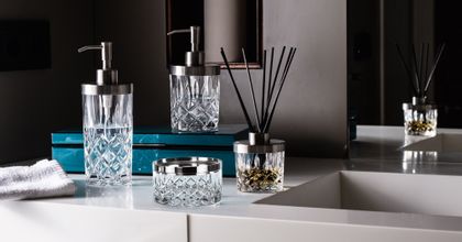 The NACHTMANN Spa Noblesse series on a bathroom sink in front of a mirror. The scent diffuser is filled with fragrance and black sticks. The small and the tall soap dispenser are filled with clear liquid. The storage jar is empty.<br/>