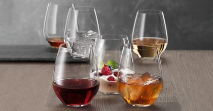 Six gobelets SPIEGELAU Authentis Casual sur une table en bois, chacun rempli différemment. Avec du vin rouge, avec une boisson rafraîchissante sur glace, avec un dessert crémeux, avec du vin blanc, avec de l'eau et des glaçons et avec une liqueur brune.<br/>