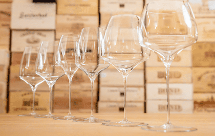 A close-up view of a line of RIEDEL wine glasses arranged in ascending size on a wooden surface, with a backdrop of wooden wine crates highlighting craftsmanship and elegance.