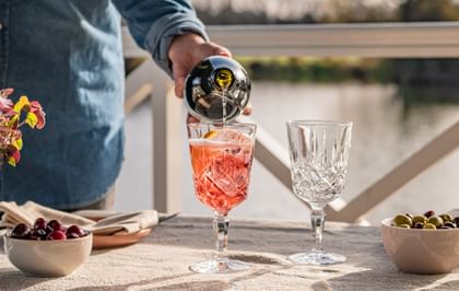Two NACHTMANN cocktail/wine glasses on a table by the lake. One of the glasses is being filled with a spritz cocktail over ice.