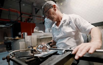 A master glassblower shapes molten glass using traditional tools in the RIEDEL glassworks, wearing a white polo shirt with the RIEDEL logo.