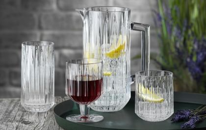 The NACHTMANN Jules crystal glass series on a round serving tray. The pitcher and the tumbler are filled with water, ice cubes and lemon slices, to their right is lavender. The goblet is filled with red wine. The longdrink glass in the background is empty.<br/>