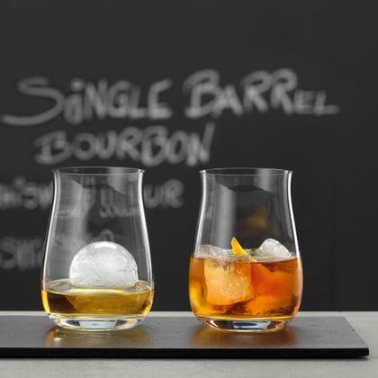 Two SPIEGELAU stemless Bourbon glasses, filled with whiskey and ice cubes. In the background a blackboard with Single Barrel Bourbon written on it.<br/>