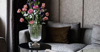 Il vaso Minerva di NACHTMANN con piede rivestito in platino e un grande bouquet di fiori su un tavolino rotondo. Sullo sfondo un divano grigio con un cuscino marrone.<br/>