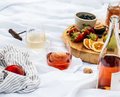 RIEDEL O Wine Tumblers filled with white wine and rose on a picnic rug. A cheese platter is on the right and a bag and wine cork on the left.