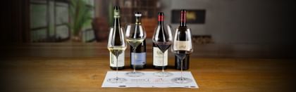 Tasting mat with 2 filled different white wine glasses and two filled different red wine glasses of the RIEDEL Veloce series in the background 2 different white wine bottles and two different red wine bottles