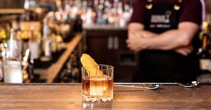 neat glass filled with a cocktail garnished with a zest, on the bar, in the background the bartender with clasped hands