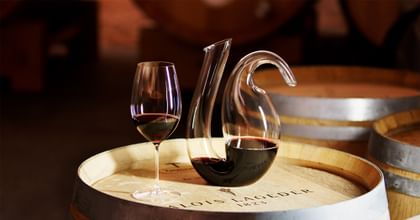 RIEDEL wine glass and RIEDEL Ayam Decanter filled with red wine on top of a wine barrel.