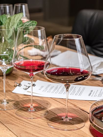 Il bicchiere SPIEGELAU Definition Burgundy pieno di vino rosso e il bicchiere Bordeaux su un tavolo di legno con un bicchiere Universal vuoto sullo sfondo.<br/>