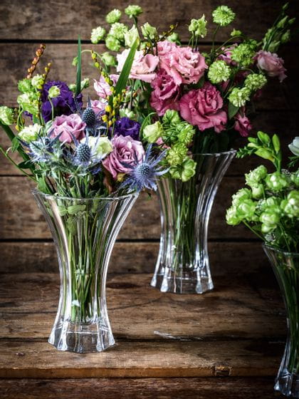 Les vases en cristal NACHTMANN Saphir, remplis de bouquets de fleurs colorés en rouge, vert, violet et bleu sur un buffet en bois.<br/>