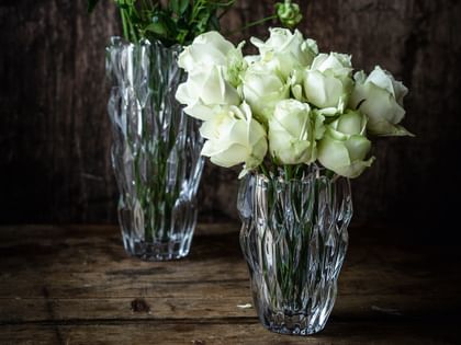 Le petit vase ovale NACHTMANN Quartz rempli de roses blanches devant le grand vase NACHTMANN Quartz rempli de fleurs blanches sur un buffet en bois foncé.<br/>