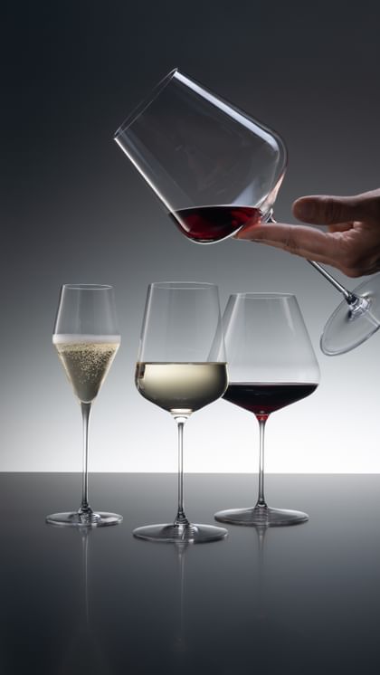 Three SPIEGELAU Definition glasses of which the Champagne glass is filled with sparkling wine, the Universal glass with white wine and the Burgundy glass with red wine. Coming from the top right corner, a hand is holding the Bordeaux glass filled with a little red wine.<br/>