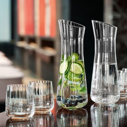 A bar counter with two SPIEGELAU Lifestyle carafes. One of them filled with infused water with blueberries, cucumber and a mint sprig, and one of the pure water. In front and behind the carafes are empty Lifestyle tumblers.<br/>