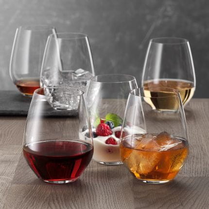 Six SPIEGELAU Authentis Casual tumblers on a wooden table, each of them filled differently. With red wine, with a soft drink on ice, with a creamy desssert, with white wine, with water and ice cubes and with a brown liquor.<br/>