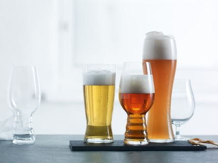 Quattro bicchieri SPIEGELAU Beer Classics in fila, uno vuoto e gli altri riempiti con birre diverse. I tre bicchieri pieni si trovano su un piatto da portata nero su uno sfondo chiaro.<br/>
