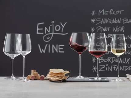 Bicchieri SPIEGELAU Willsberger Anniversary su una credenza di marmo. A sinistra un bicchiere da Bordeaux e un bicchiere da vino bianco vuoti, al centro una pila di tappi di sughero e patatine, a destra il bicchiere da Borgogna pieno, il bicchiere da vino rosso pieno e il bicchiere da vino bianco pieno. Sullo sfondo una lavagna con un menu e la scritta bere vino.<br/>