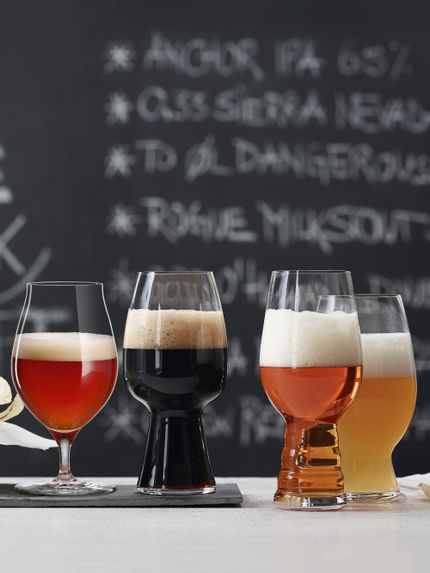 The with beer filled SPIEGELAU Craft Beer Glasses on a table in front of a blackboard with a menu. The glass for Barrel Aged beer and the Stout glass are standing on a slate plate, the IPA glass and the American Wheat Beer glass next to it on the table.<br/>