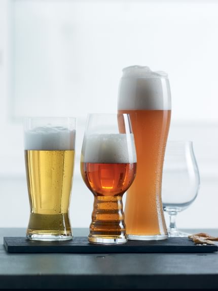 Four SPIEGELAU Beer Classics glasses in a row one is empty and the rest filled with different beers. The three filled glasses are on a black serving platter against a light background.<br/>