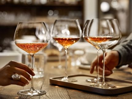 A hand holding the SPIEGELAU Burgundy glass of the series Willsberger Anniversary, filled with rosé colored sparkling wine. Further back on the wooden table, there are more glasses.<br/>