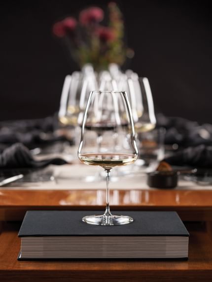 Single RIEDEL Winewings glass filled on set table elevated on a grey book. Behind is other RIEDEL Winewings glasses filled.