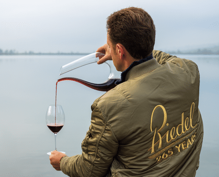 Max Riedel pouring wine from decanter into wine glass with a lake in the background.