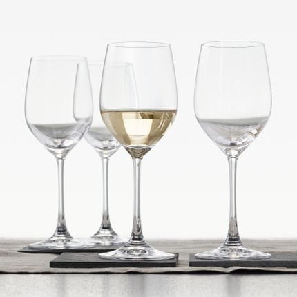 Cuatro copas de vino blanco SPIEGELAU Vino Grande sobre una mesa con mantel. Las dos copas en primer plano están sobre posavasos de pizarra, una de ellas llena de vino blanco.<br/>