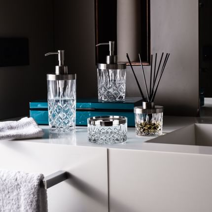 The NACHTMANN Spa Noblesse series on a bathroom sink in front of a mirror. The scent diffuser is filled with fragrance and black sticks. The small and the tall soap dispenser are filled with clear liquid. The storage jar is empty.<br/>