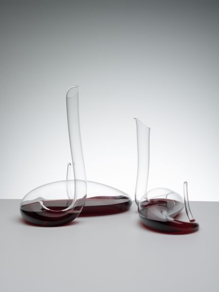 A RIEDEL Mamba Decanter on the left and a RIEDEL Mamba Mini Decanter on the right. Both are filled with red wine on a grey background.