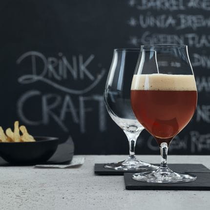 Two SPIEGELAU Craft Beer Glasses for Barrel Aged Beer on slate trays. The glass in the foreground is filled with Barrel Aged Beer, in the background are potato chips and a blackboard with a beer menu on it.<br/>
