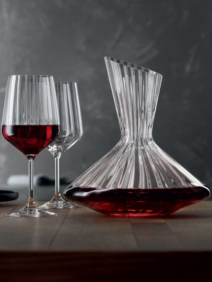The with red wine filled SPIEGELAU Lifestyle decanter on a table. Left of the decanter two Lifestyle red wine glasses, one of them filled with red wine. On the left edge of the table an empty black plate.<br/>