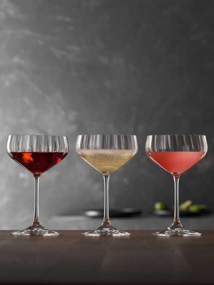 Four SPIEGELAU Lifestyle Coupette glasses on a wooden table. The glasses are filled with different cocktails. From left to right with a Manhattan, a Prosecco, a Cosmopolitan and a Margaritha.<br/>