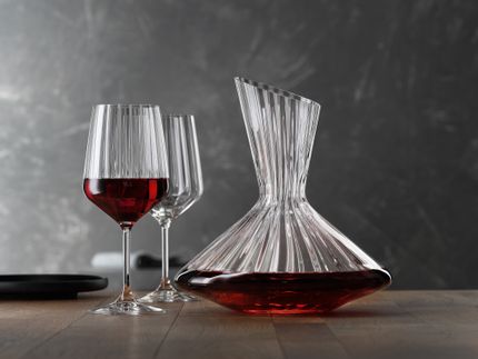The with red wine filled SPIEGELAU Lifestyle decanter on a table. Left of the decanter two Lifestyle red wine glasses, one of them filled with red wine. On the left edge of the table an empty black plate.<br/>