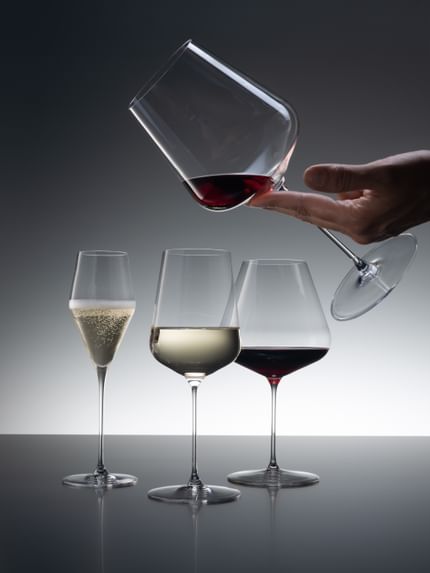 Tre bicchieri SPIEGELAU Definition di cui il bicchiere Champagne è riempito di spumante, il bicchiere Universal di vino bianco e il bicchiere Burgundy di vino rosso. Nell'angolo in alto a destra, una mano tiene il bicchiere Bordeaux riempito con un po' di vino rosso.<br/>