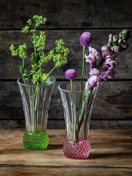 Kleine NACHTMANN-Vasen SPRING auf einem Holztisch. Eine hat einen limonenfarbenen Boden und eine grüne Blume darin, die andere hat einen roséfarbenen Boden und rosa Blumen darin.<br/>