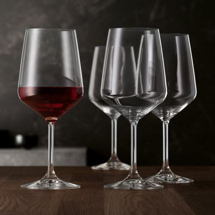 Quatre verres à vin rouge de style SPIEGELAU sur une table en bois. Un verre est rempli de vin rouge.<br/>