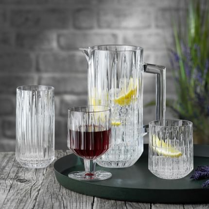 La série de verres en cristal NACHTMANN Jules sur un plateau de service rond. Le pichet et le gobelet sont remplis d'eau, de glaçons et de tranches de citron, à leur droite se trouve de la lavande. Le gobelet est rempli de vin rouge. Le verre à long drink à l'arrière-plan est vide.<br/>