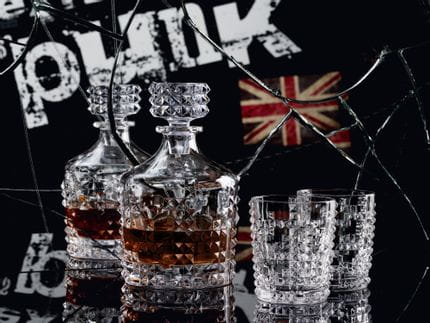 The with whisky filled NACHTMANN Punk decanter and two Punk whisky tumblers on a black table in front of a broken mirror with the written word punk and a junion jack in this mirror.<br/>
