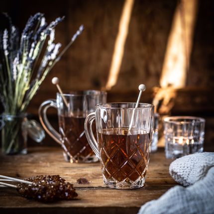 Der NACHTMANN Noblesse Teebecher mit heißem Tee und Kandiszucker. Auf dem Holztisch liegen weitere Stangen mit braunem Kandiszucker, ein Krug mit Lavendelblüten, ein zweiter gefüllter Kristallteebecher und ein leuchtendes Teelicht.<br/>