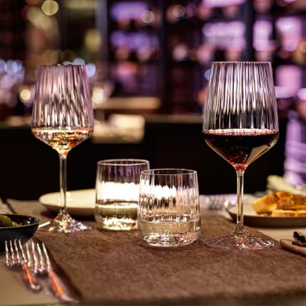 La serie SPIEGELAU Lifestyle sul tavolo di un ristorante in un'accogliente atmosfera serale. La luce viola risplende attraverso la struttura ondulata del bicchiere da vino bianco, del bicchiere da vino rosso, dei bicchieri da acqua e della caraffa.<br/>