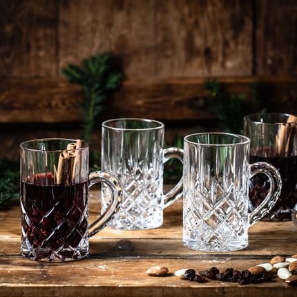 El vaso de té NACHTMANN Noblesse con vino caliente y canela en rama sobre un aparador de madera. A su lado hay dos vasos de té Noblesse vacíos y uno lleno, almendras, pasas y ramas de pino.<br/>