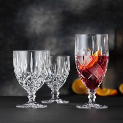 Le verre à boisson glacée Noblesse de NACHTMANN rempli d'une boisson cocktail rouge, de glaçons et d'une tranche d'orange à côté du grand et du petit gobelet vides. En arrière-plan, des oranges.<br/>