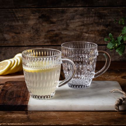 Two NACHTMANN Ethno hot beverage mugs on a cutting board. One mug is filled with a hot lemon drink, the one behind is empty. In the background are lemon slices.<br/>